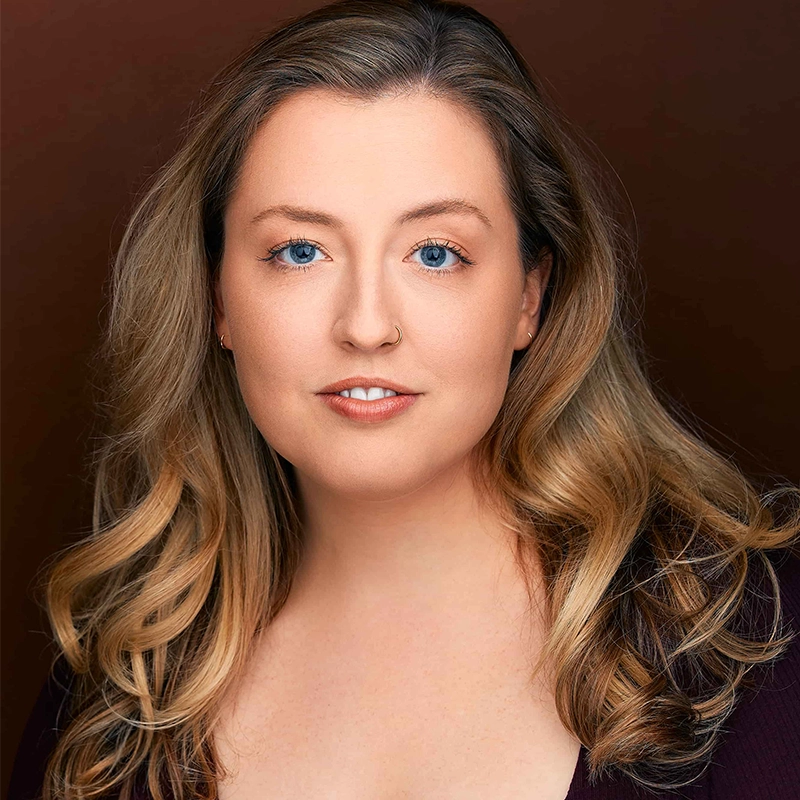 Chris Allyah, a woman with wavy blonde hair and blue eyes, is shown against a dark brown background. She has fair skin, natural makeup, and wears a dark top, looking directly at the camera with a neutral, slight smile.
