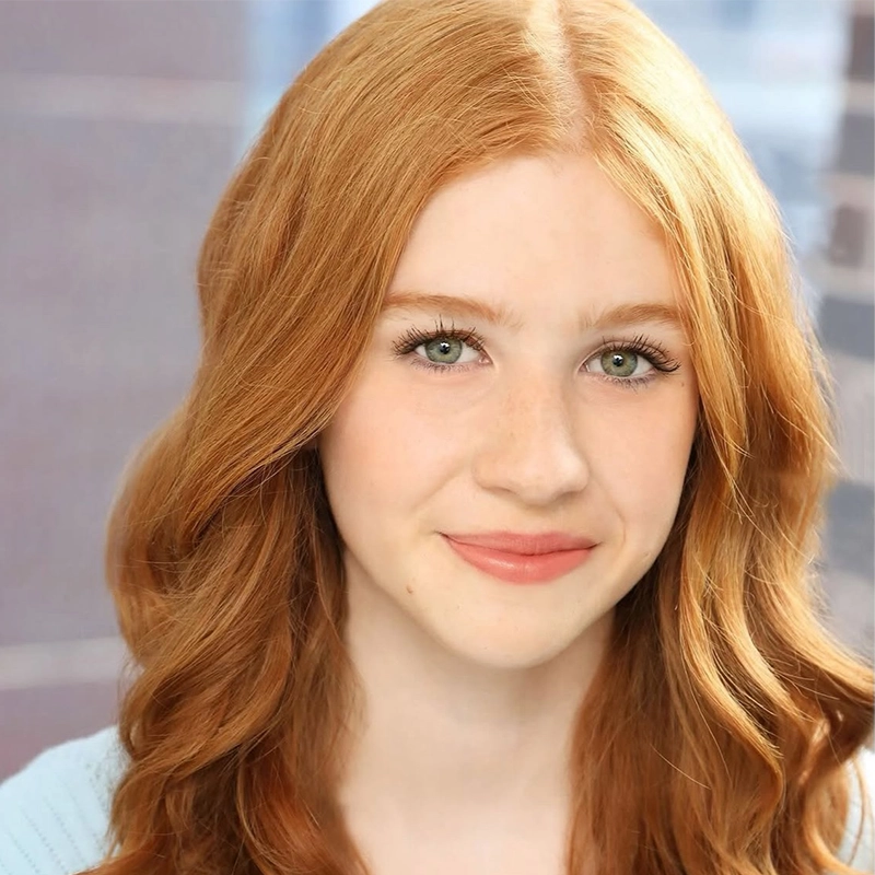 A young person with long, wavy red hair and fair skin smiles gently. They have green eyes, light makeup, and are wearing a pale blue top. The softly blurred background with cool tones highlights their face in this Matthew Brand-inspired portrait.