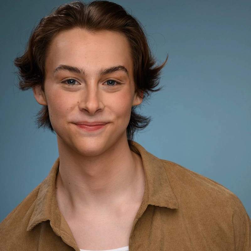 A young person with light skin, medium brown hair, and blue eyes is smiling softly. They wear a tan corduroy jacket over a white shirt, with a blue background behind them. Their hair is parted down the middle and slightly tousled.