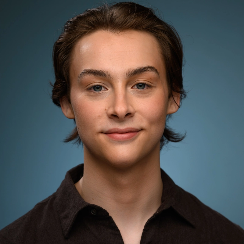 A young person with fair skin, straight brown hair parted in the middle, and blue eyes is smiling softly. They are wearing a dark button-up shirt and are posed in front of a plain blue gradient background.