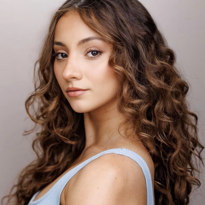 A young woman with long, voluminous brown curly hair poses against a light background. With soft makeup and brown eyes, Julia Grondin wears a light blue sleeveless top, calmly looking over her shoulder at the camera.