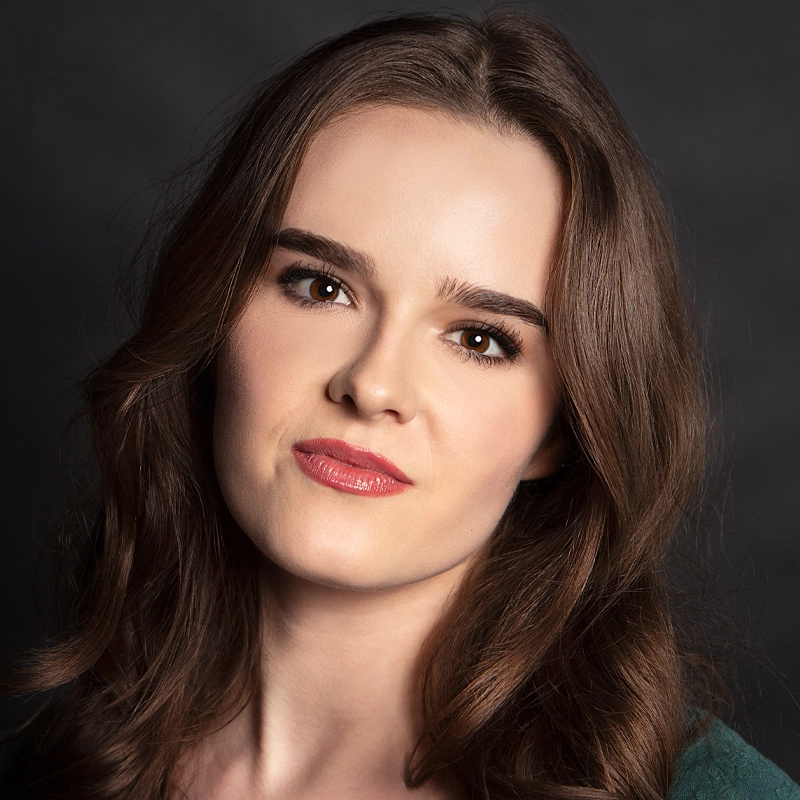Julietta Guinasso, a young woman with long, wavy brown hair, fair skin, and brown eyes, looks at the camera with a neutral expression. She wears natural makeup and a green top against a dark, softly lit background.