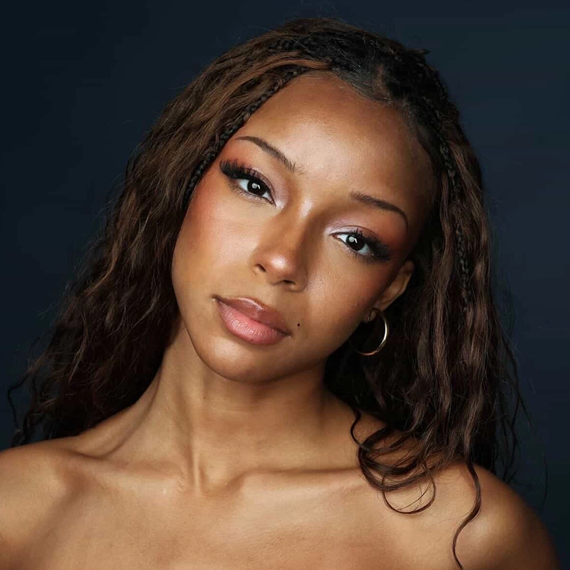 A young woman, reminiscent of Hailey Morales, with long, wavy brown hair and subtle braids poses against a dark background. She wears hoop earrings and natural makeup with warm tones and glossy lips, softly gazing into the camera.