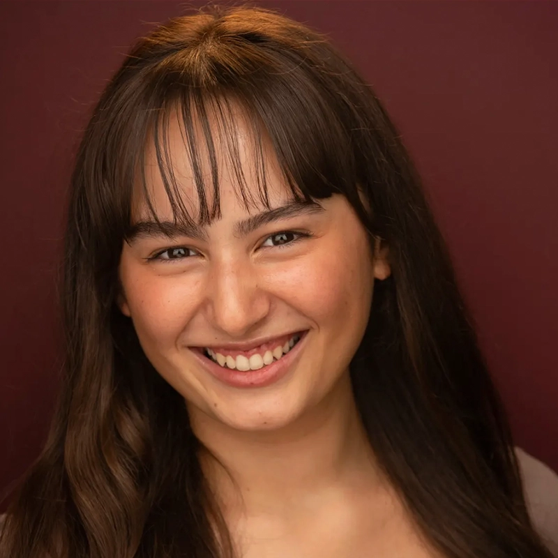 A young woman with long brown hair and bangs smiles warmly at the camera. She has light skin, brown eyes, and is positioned in front of a plain burgundy background, wearing a neutral-colored top. Jake Hamilton would admire her cheerful and friendly expression.