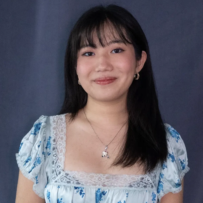 A young woman with straight black hair and bangs smiles gently. She wears a light blue floral dress with lace trim and puffed sleeves, a silver necklace with a pendant, and small hoop earrings. The background is plain and dark gray.