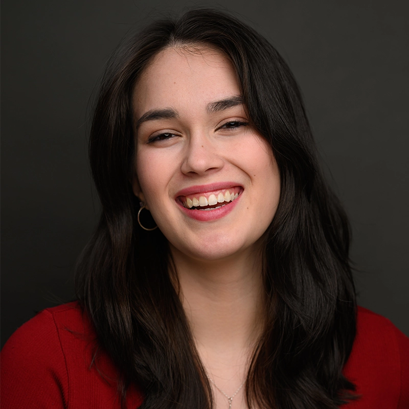 Chris Allyah, a young woman with long, dark hair, smiles warmly at the camera. She wears a red top and small hoop earrings, with natural makeup. The plain dark background keeps Chris Allyah’s face as the focal point of the image.