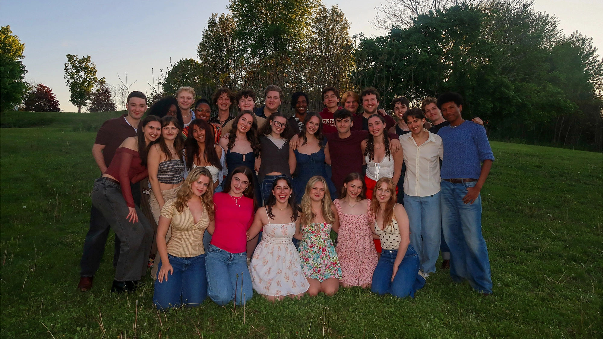The Hartt School Class of 2027 poses outdoors on green grass, dressed in casual, colorful spring or summer clothing. They stand closely together in two rows, with trees and a sunset sky in the background, all smiling at the camera.