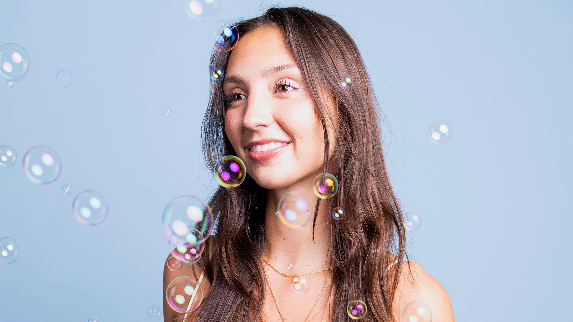 A young woman with long brown hair smiles softly, looking off camera against a light blue background. She wears layered gold necklaces and a sleeveless top. Iridescent soap bubbles float around her, evoking the whimsical charm of Bubbling Cabaret.