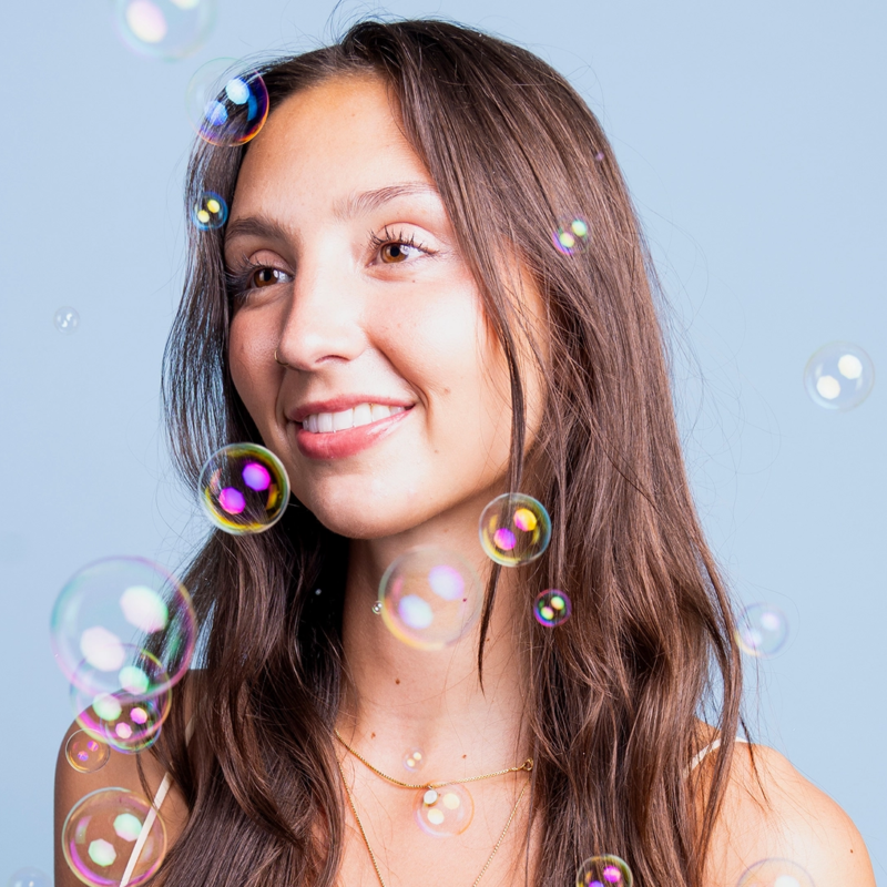 A young woman with long brown hair smiles softly, looking slightly to the side. She wears a light top and gold necklaces. Transparent soap bubbles float around her against a solid light blue background, creating a playful and dreamy atmosphere.