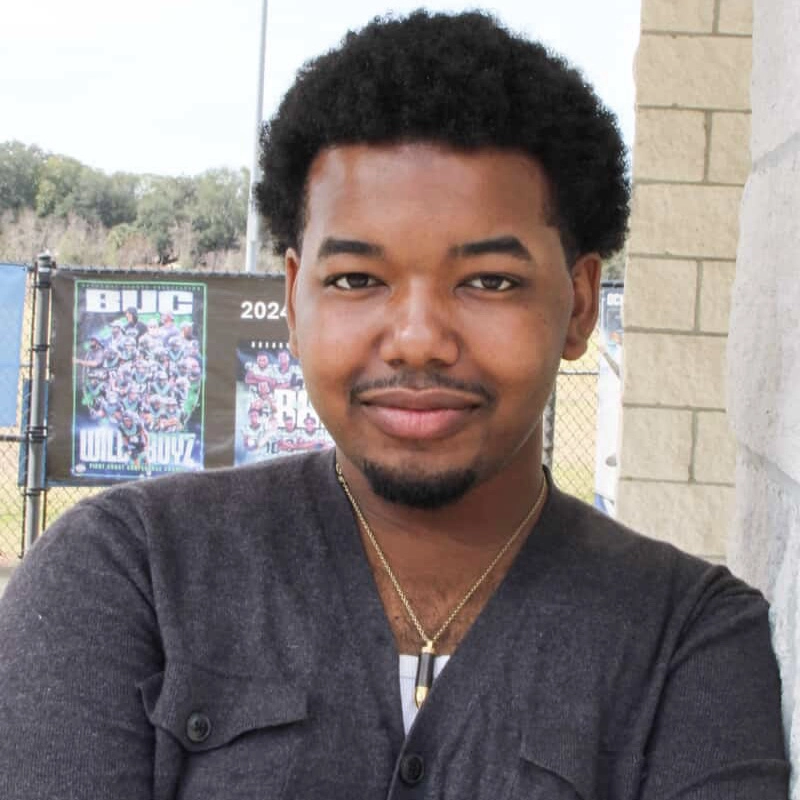 A young man with short curly hair and a trimmed beard stands smiling with arms crossed. Israel Haynes is wearing a gray buttoned sweater and a gold necklace, framed by sports posters on a chain-link fence and outdoor trees.