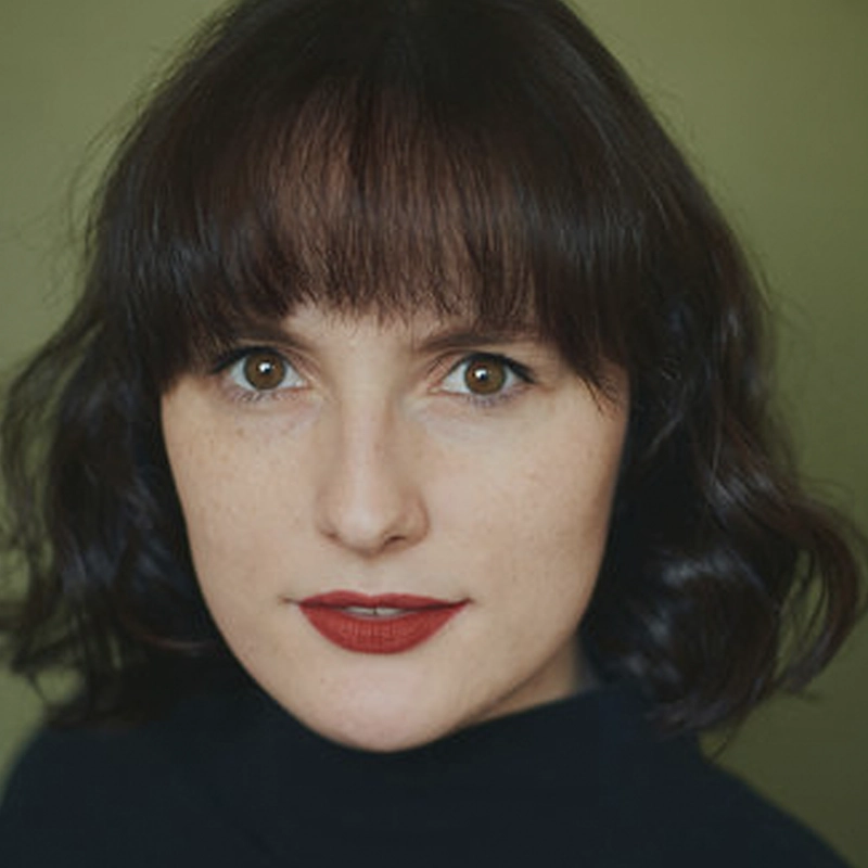 Santa Claire Hirsch, a woman with wavy dark brown hair and bangs, looks directly at the camera. She has fair skin, brown eyes, and wears bright red lipstick. She is dressed in a dark top against a plain olive-green background.