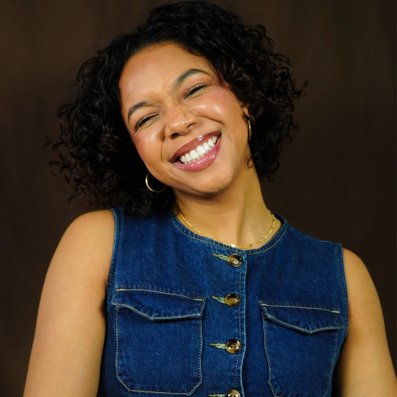 A smiling woman with curly hair stands against a dark brown background. She is wearing a sleeveless denim top with gold buttons and hoop earrings, tilting her head slightly and squinting her eyes cheerfully.