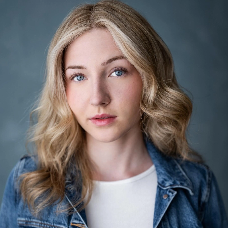 A young woman with wavy blonde hair and blue eyes looks into the camera. She wears a white top under a blue denim jacket. The background is a soft, muted gray. Her expression is neutral, and her hair frames her face, embodying the calm confidence often associated with Emersyn Hunt.