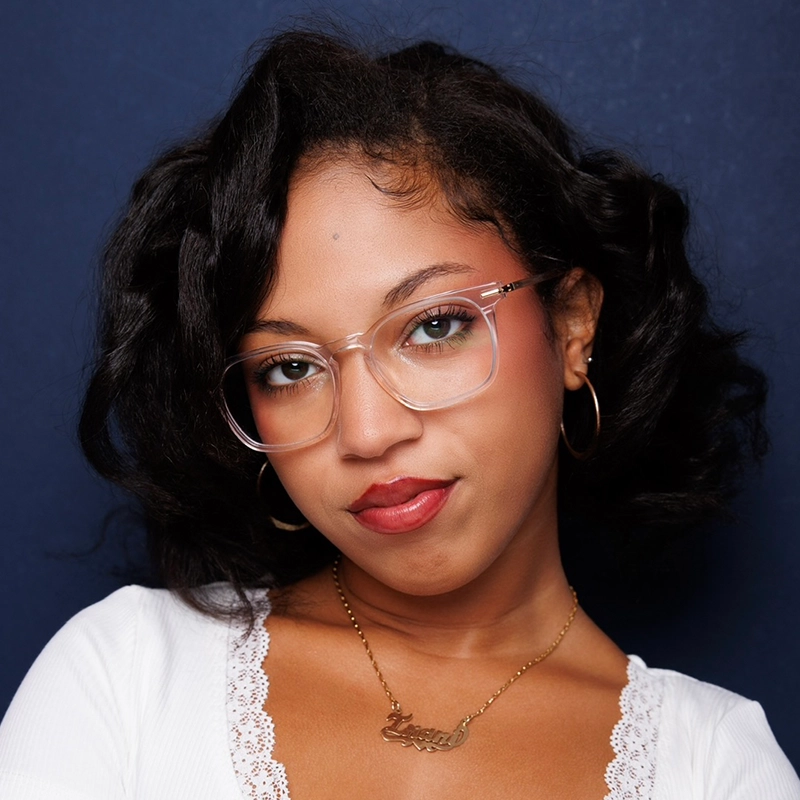 A woman with wavy black hair, clear-framed glasses, and hoop earrings stands against a dark blue background. She wears a white top with lace trim and a gold necklace. She gazes confidently at the camera with a slight smile.
