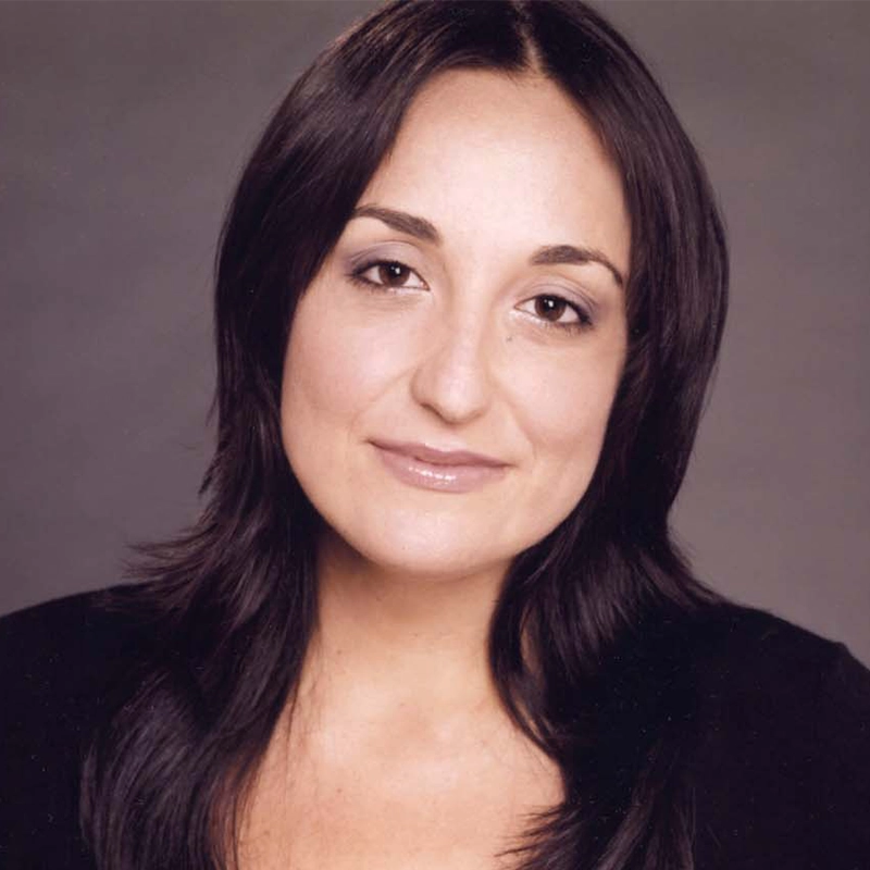A woman with long, dark brown hair and fair skin smiles softly at the camera. She has brown eyes, natural makeup, and wears a black top. The background is plain and gray, with soft lighting highlighting her face.