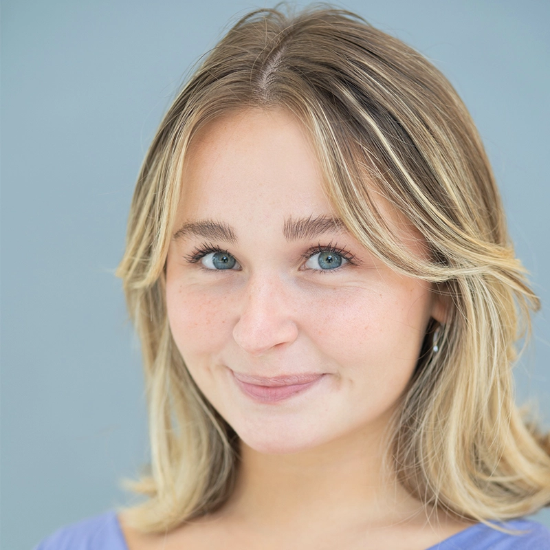 A young woman with shoulder-length blonde hair and blue eyes smiles slightly at the camera. She is wearing a light purple top and standing against a plain light blue background. Her expression is friendly and calm.