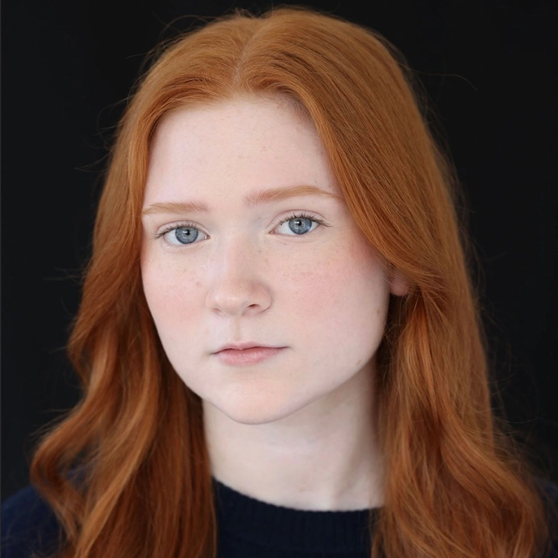A young woman with long, wavy red hair and fair skin stares calmly at the camera. She has light blue eyes, light brows, and a neutral expression. She is wearing a dark top, and the background is solid black.