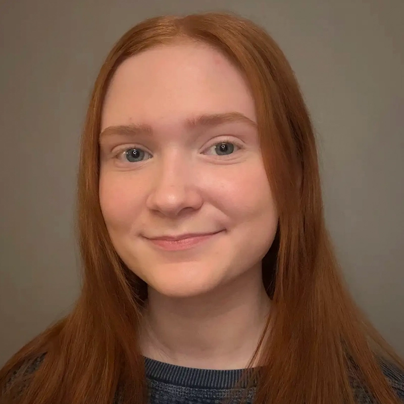 A young person with straight, long red hair and fair skin smiles gently. Jake Hamilton has light-colored eyes and wears a dark, textured sweater. The plain, neutral background draws focus to his face and expression.