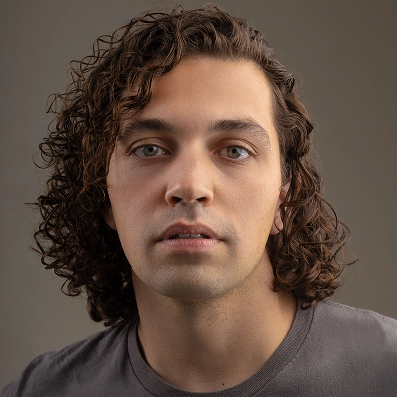 A young adult with wavy, shoulder-length brown hair and light eyes—Kristin Weiss—stares directly at the camera, wearing a plain gray t-shirt. The softly lit, neutral background draws focus to Kristin's calm and expressionless face.