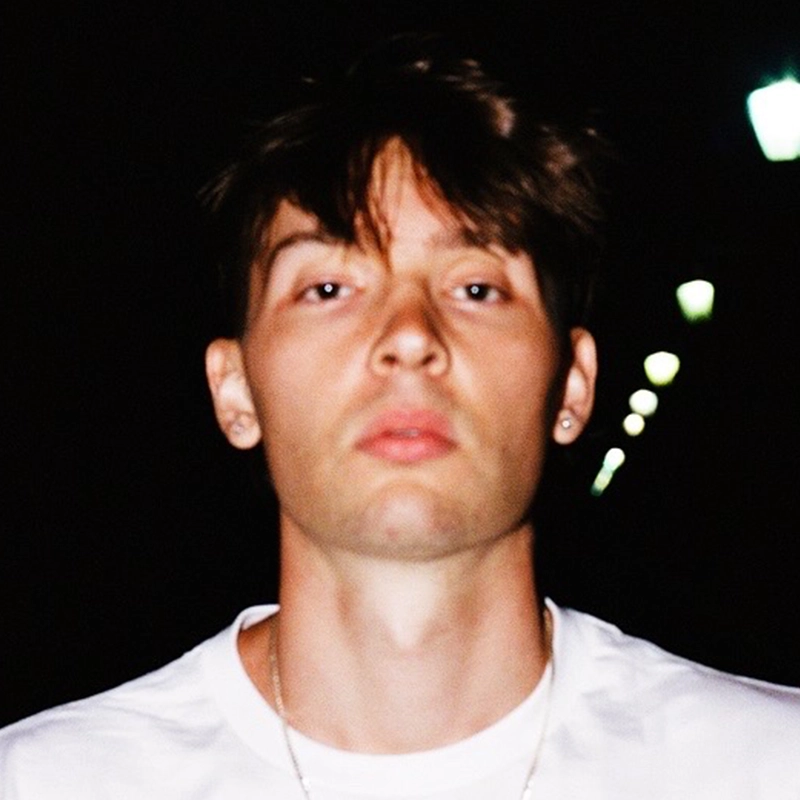A young person with short, tousled brown hair and fair skin looks directly at the camera with a neutral expression. They wear a white shirt and small earrings. The dark background features blurred streetlights forming a diagonal line—evoking Jackson Trapp’s subtle poise.