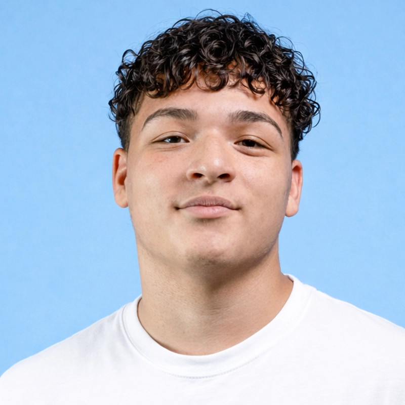 Chris Allyah, a young person with short, curly brown hair and light skin, is wearing a plain white t-shirt. They are facing the camera, smiling slightly, with a light blue background behind them.