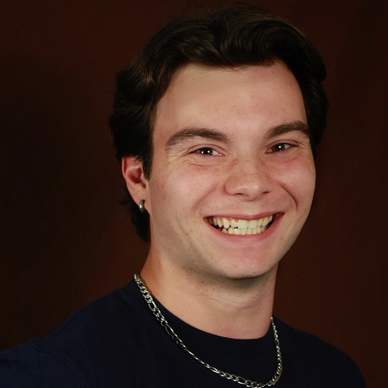 Chris Allyah, a young man with short brown hair, smiles widely, showing his teeth. He wears a dark shirt, a silver chain necklace, and a small hoop earring in his left ear. The dark brown background and lighting highlight his face.