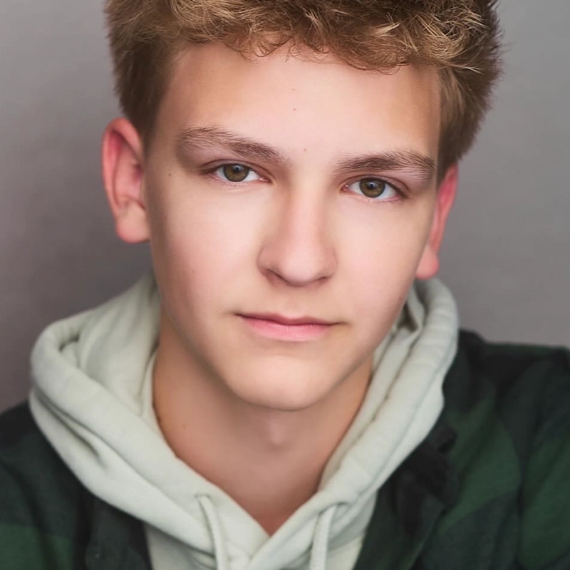 Jake Hamilton, a teenage boy with short, light brown hair and hazel eyes, looks at the camera with a neutral expression. He is wearing a light gray hoodie under a dark green and black plaid shirt against a softly lit plain background.