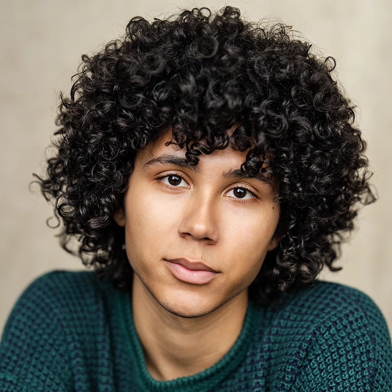 Chris Allyah, with medium skin tone and dark, curly hair, looks softly at the camera. They wear a green textured sweater. The background is neutral and blurred, highlighting their calm expression.