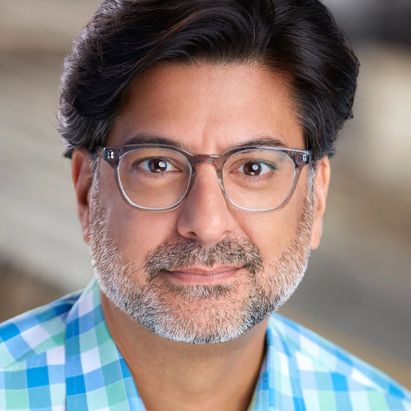 A middle-aged man with short, wavy dark hair and gray streaks, sporting a salt-and-pepper beard and mustache. He wears clear-framed glasses and a blue-green plaid shirt, looking at the camera with a slight smile against a blurred background.