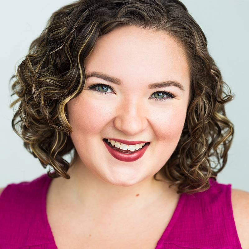 A young woman, Jenna Leigh Miller, with short, curly brown hair and light skin smiles brightly. She wears a sleeveless magenta top and has natural makeup with bold lipstick. The plain, light background highlights her cheerful expression.