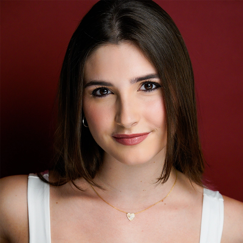 Chris Allyah, a young woman with straight brown hair and brown eyes, smiles softly at the camera. She wears a white top, a gold necklace with a heart pendant, and subtle makeup against a solid dark red background.