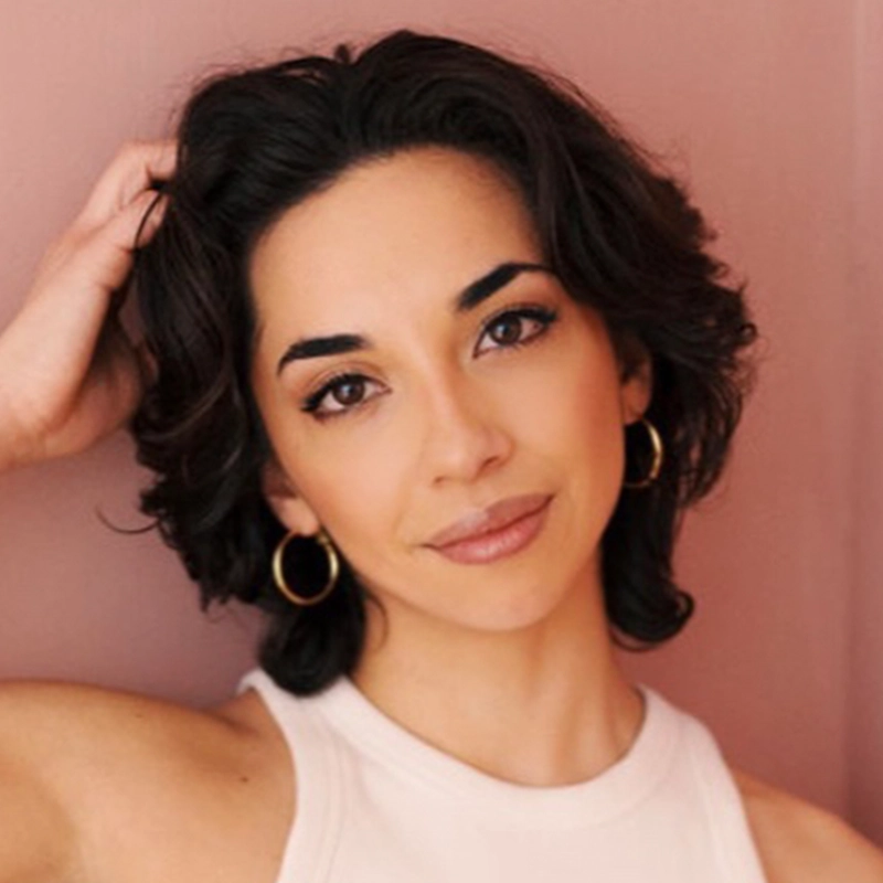 Sandra Valls, with wavy, dark brown, chin-length hair, gold hoop earrings, and a sleeveless white top, poses against a soft pink background. She has natural makeup and a subtle smile, with one hand gently touching her hair.