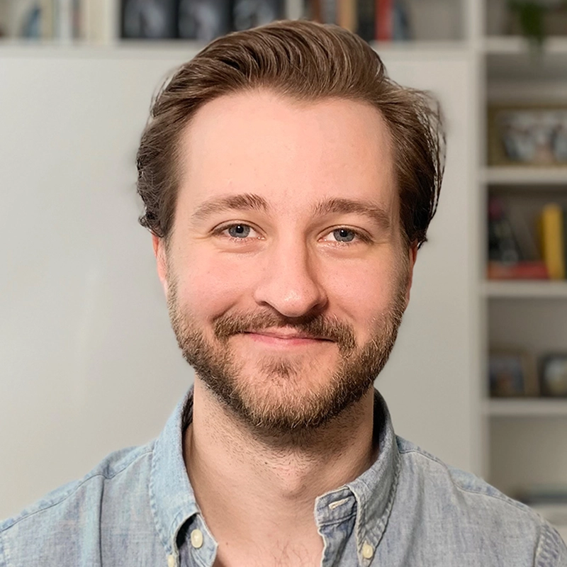 A smiling man with light skin, short brown hair, and a trimmed beard faces the camera. He wears a light blue button-down shirt. The background is softly focused, showing shelves with books and small decor items.