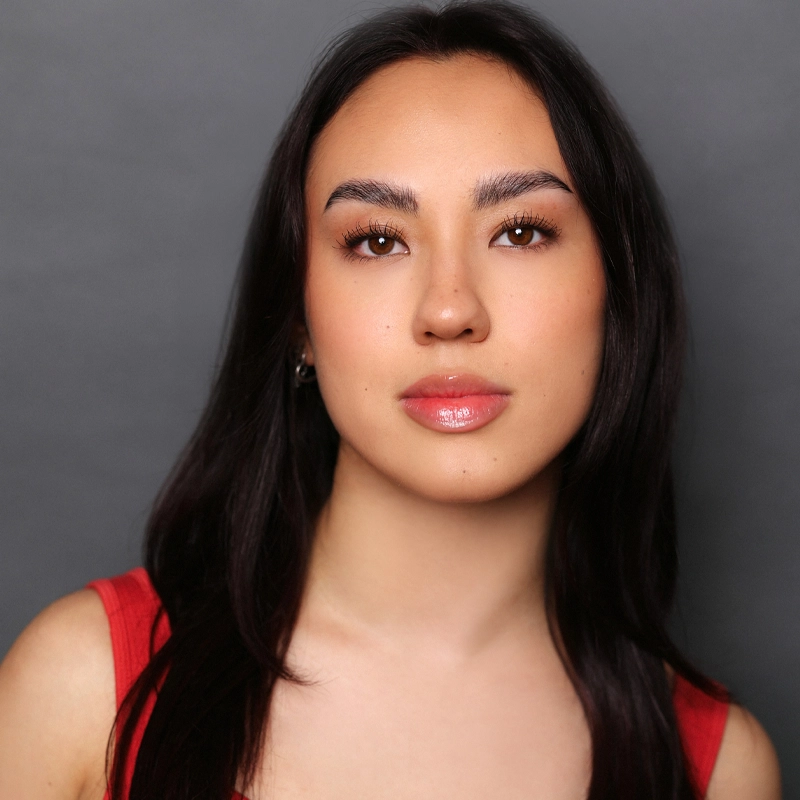 Sydney Jones, a young woman with long dark hair, gazes calmly at the camera. She wears a sleeveless red top, has natural makeup with glossy lips, and a soft complexion. The plain dark gray background creates a professional portrait look.
