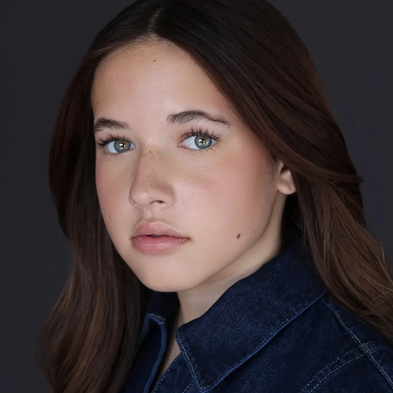 Jordan Gulliford, a young person with long brown hair and green eyes, gazes seriously at the camera. With fair skin, light makeup, and a small beauty mark on their cheek, they wear a dark denim jacket against a plain dark gray background.