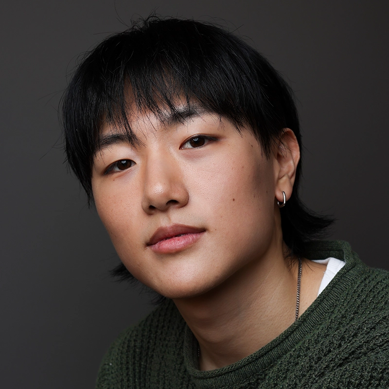 A young person with straight black hair and an earring looks into the camera with a calm expression. They are wearing a green sweater over a white shirt. The background is plain and dark, putting focus on their face.