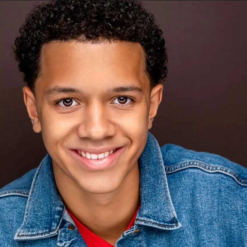 A young person with short, curly hair smiles at the camera. Jordan Gulliford wears a blue denim jacket over a red shirt and is posed against a dark, solid background. The lighting highlights their friendly and approachable expression.