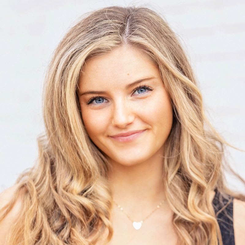 Chris Allyah, a young woman with long, wavy blonde hair and blue eyes, smiles softly. She wears a black top and a gold necklace with a heart-shaped pendant. The light, out-of-focus background draws attention to her face.