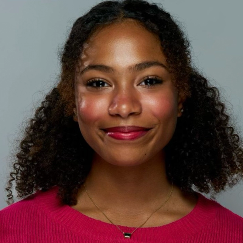 A young woman with curly dark hair smiles warmly. Jordan Gulliford is wearing a bright pink sweater and a delicate gold necklace. Her skin is glowing, with rosy cheeks, well-defined brows, and natural makeup against a plain light gray background.
