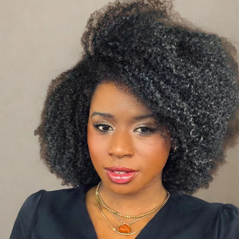 A woman with voluminous, natural curly black hair poses against a plain background. She wears a black top, layered gold necklaces, and natural makeup with shimmery eye shadow, winged liner, and glossy red lips. She looks confidently at the camera.