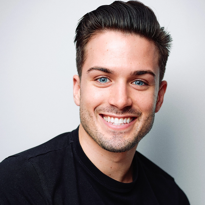 A young man with short, neatly styled dark brown hair and light skin smiles at the camera. He has blue eyes, trimmed facial hair, and wears a black crew-neck shirt. The background is a plain, light grey.