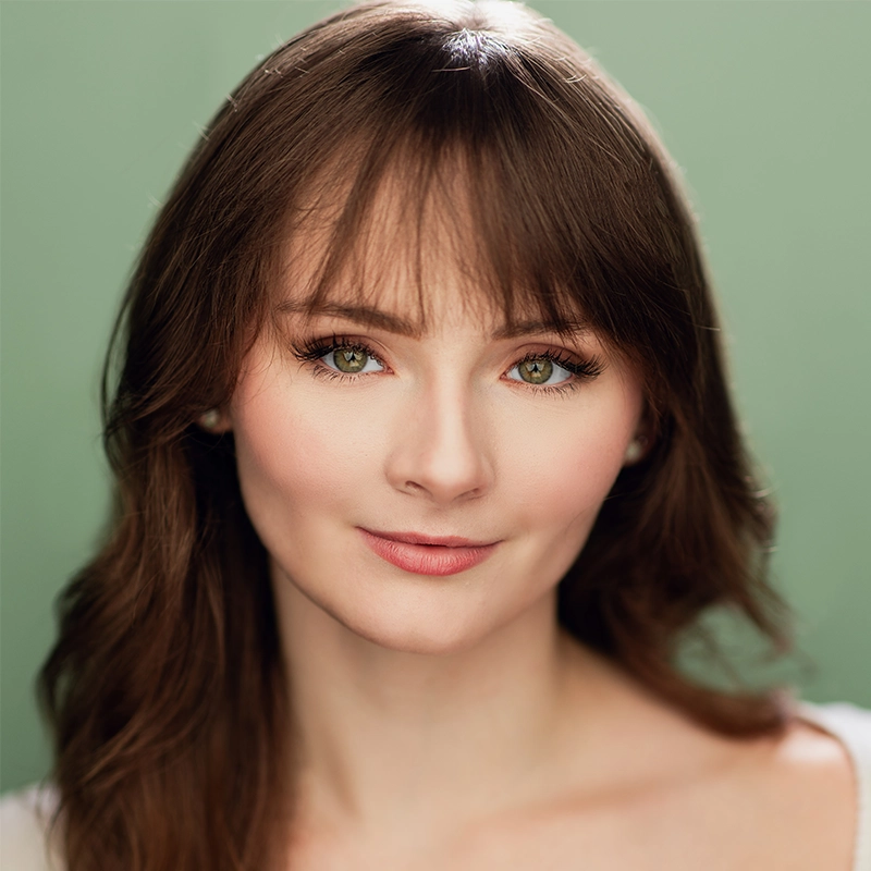 A young woman with fair skin, straight brown hair, and bangs smiles softly. She has green eyes, natural makeup, and is wearing a white top. The soft green background gently contrasts with her hair and complexion—her name is Katharine Yeargin.