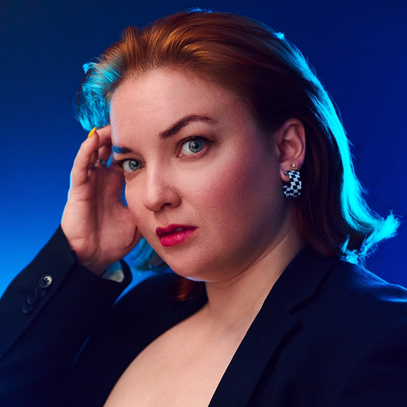 A woman with fair skin and auburn hair looks at the camera with a serious expression. She wears a black blazer, bold pink lipstick, and checkerboard-patterned earrings. Her hand touches her temple. The background is illuminated with blue light.