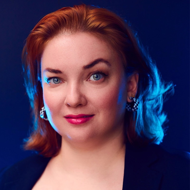 A woman with fair skin and shoulder-length reddish hair styled back, resembling Sandra Valls, wears a dark blazer and sparkly earrings. She has blue eyes, pink lipstick, and a confident expression against a dark blue background with blue lighting.