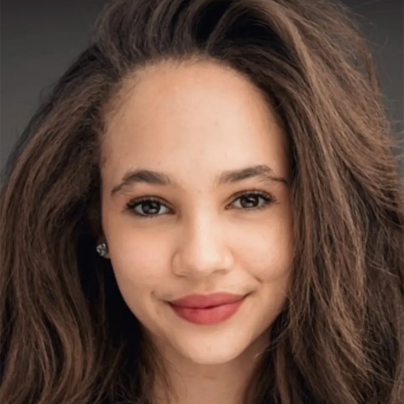 A young woman with long, voluminous brown hair smiles softly at the camera. She has light skin, brown eyes, and natural makeup with rosy lips. Jordan Gulliford wears stud earrings; the plain dark background keeps the focus on her face.