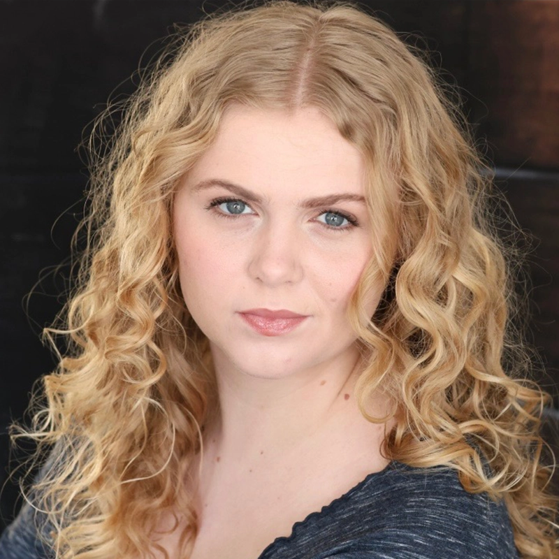 Maya Monteleone, a woman with long, curly blonde hair and fair skin, looks directly at the camera with a neutral expression. Her blue eyes and dark top stand out against the dark, blurred background that highlights her face.