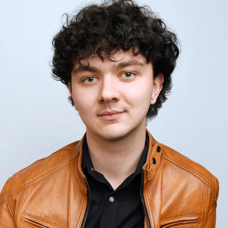 Ryan P. Kennedy, a young person with curly dark hair and fair skin, wears a light brown leather jacket over a black collared shirt. Light hazel eyes and a slight smile complement the look, with a small earring in their left ear against a plain background.