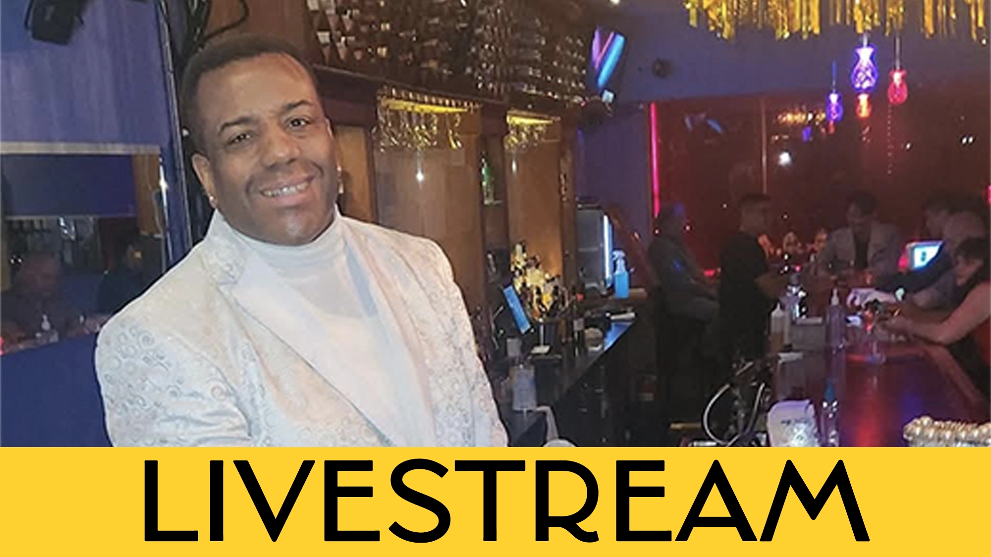 A smiling Kenny Allan Smith in a white patterned jacket stands by a bar in a lively, colorful nightclub. Behind him, people are seated at the bar with neon lights glowing. 