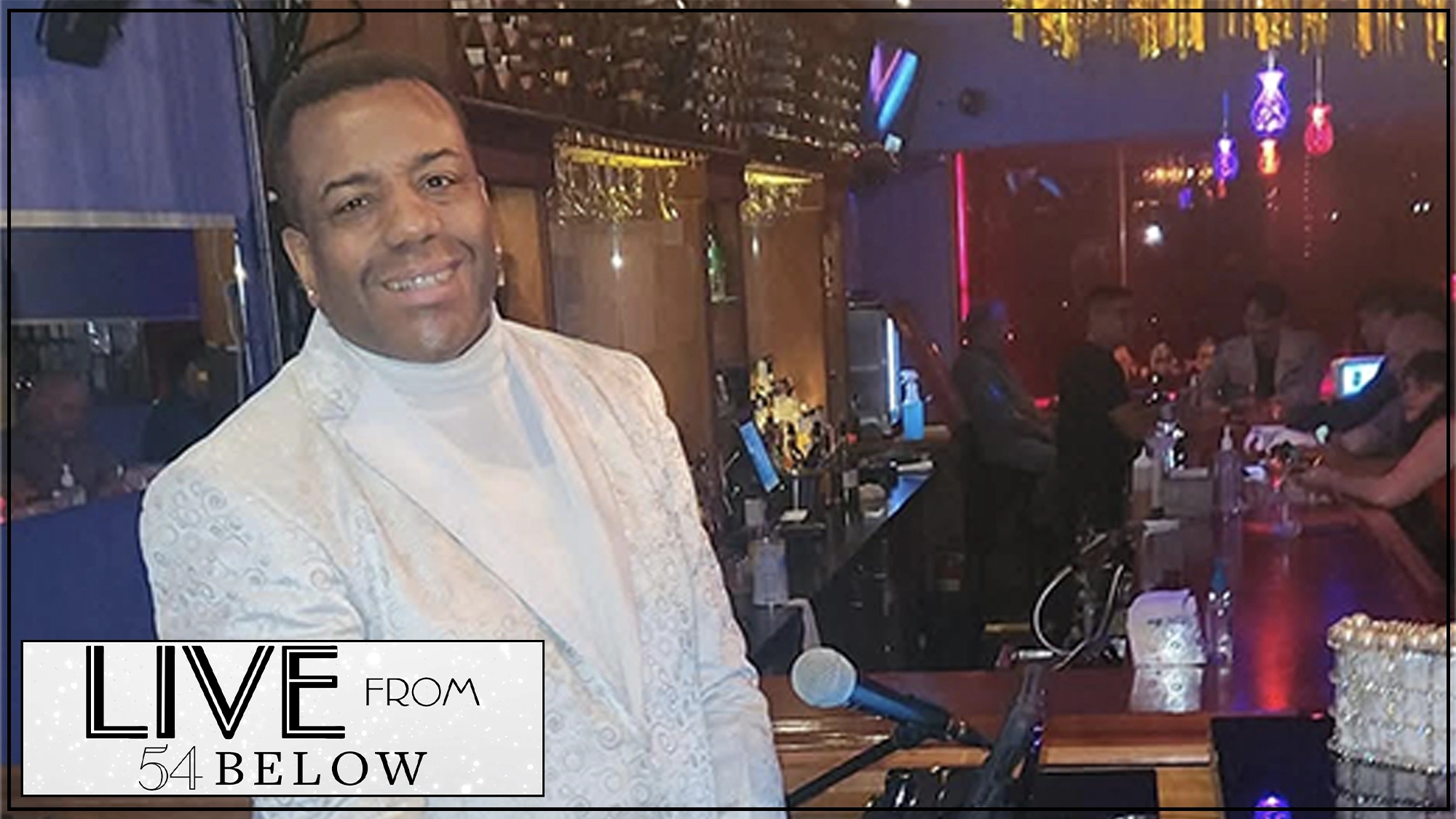 A smiling man in a white patterned suit stands at a grand piano with a microphone in front of him, inside a warmly lit bar. People are seated at the bar in the background. In the corner, the logo reads “LIVE FROM 54 BELOW: Kenny’s Turn Livestream.”.