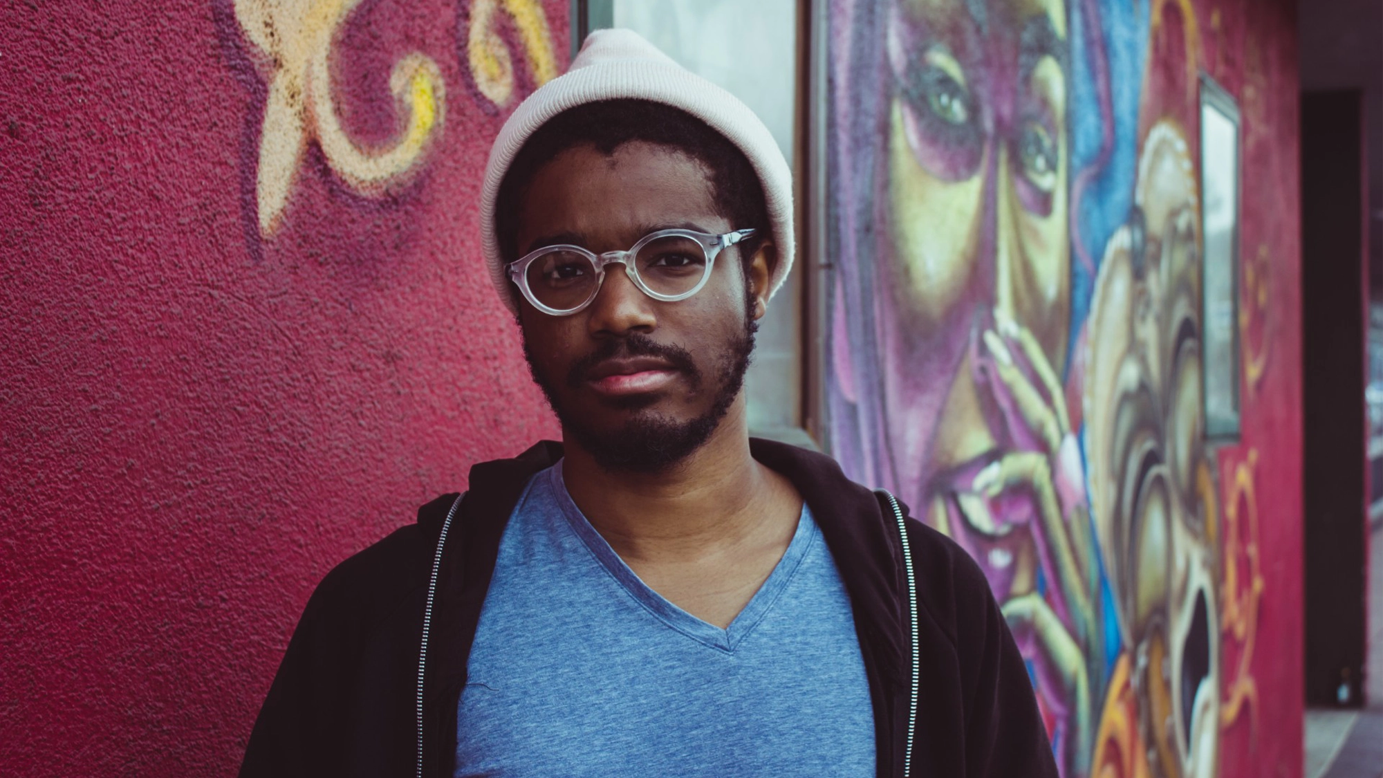 A man wearing glasses, a white beanie, and a blue V-neck shirt stands in front of a vibrant graffiti wall featuring colorful, abstract faces. He has a neutral expression and a short beard. The striking street art background evokes the dynamic energy of live music, as if the scene captures the spirit of an urban concert—perhaps even one by Khiyon.